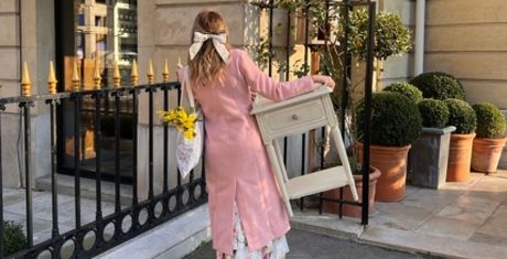 Woman in pink coat holding a chair and flowers, walking on a city street with potted plants and a decorative fence.