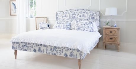 Elegant bedroom with a bed featuring blue floral bedding, a wooden nightstand with a lamp, and a bright window with white curtains.
