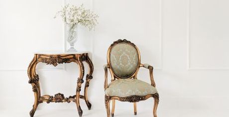 Elegant vintage chair and ornate table with a vase of white flowers against a white wall.