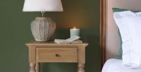 A wooden bedside table with a lamp, an open book, a lit candle, and a bed with white and green bedding against a dark green wall.