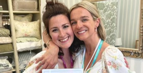 Two women smiling and embracing in a cozy room with patterned pillows and a mirror in the background.