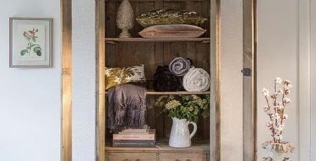 Wooden cabinet with open doors displaying pillows, rolled blankets, books, and a white jug with flowers. Framed art hangs on the white wall.