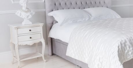 Elegant bedroom with a tufted gray headboard, white bedding, a classic white nightstand, and a bust sculpture on top.