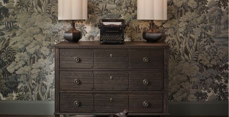 Vintage typewriter on a dark wooden dresser with two lamps, set against a detailed forest-themed wallpaper.