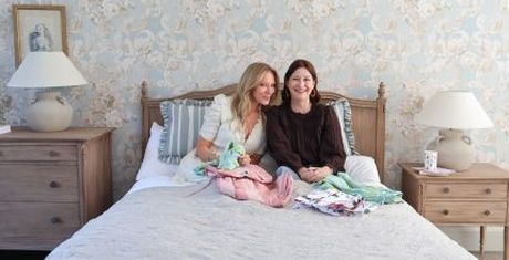 Two women sitting on a bed with floral wallpaper and wooden nightstands, surrounded by colorful clothes.