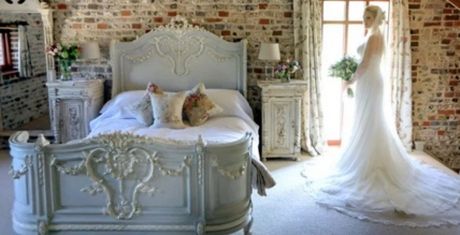 A bride in a white gown stands by a window in a rustic bedroom with an ornate bed and exposed brick walls, holding a bouquet.