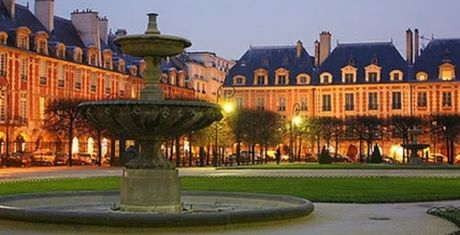 Illuminated historic square with a central fountain, surrounded by elegant, symmetrical buildings at dusk.