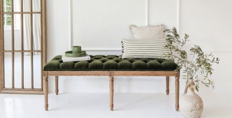 Elegant room with a green tufted bench, striped and textured pillows, a potted plant, and a tall wooden-framed mirror against a white wall.