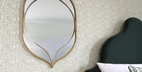 Elegant, leaf-shaped mirror with a gold frame on a patterned wallpapered wall, next to a dark green upholstered headboard.