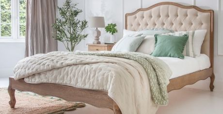 Elegant bedroom with a beige tufted headboard, green and beige bedding, a bedside table with a lamp, and a potted plant by the window.