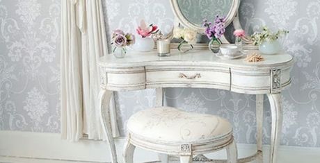 Vintage vanity table with a round mirror, floral arrangements, and a cushioned stool against a patterned wallpaper background.
