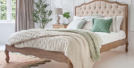 Elegant bedroom with a tufted headboard, white and green bedding, a bedside table with a lamp, a plant, and a window with curtains.