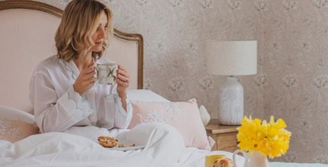 Woman in bed with a cup, wearing pajamas. Breakfast tray with pastries and a vase of yellow flowers on the bedside table.