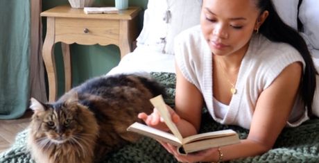 Woman reading a book on a bed with a fluffy cat lying beside her. A wooden side table and green blanket are in the background.