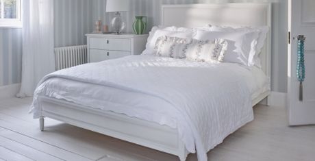 Bright bedroom with a white bed, fluffy pillows, and light blue striped walls. A lamp and decorative items sit on a white nightstand.
