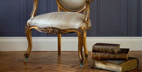 Elegant vintage chair with ornate wooden frame beside a stack of antique books on a herringbone wood floor, set against a dark paneled wall.