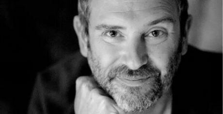 Black and white portrait of a man with a beard, smiling softly, resting his chin on his hand, and looking directly at the camera.