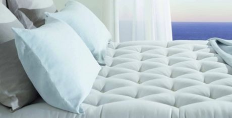 A neatly made bed with light blue pillows and a quilted white comforter, set near a window with a view of the ocean and sky.