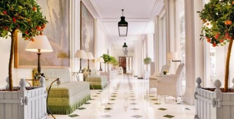 Elegant hallway with checkered marble floor, green sofas, potted orange trees, and large windows. Lantern lights hang from the ceiling.