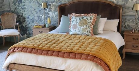 Cozy bedroom with a wooden bed, decorative pillows, a mustard yellow quilt, and floral wallpaper. A chair and nightstands flank the bed.