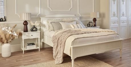 Elegant bedroom with a white bed, beige bedding, and a plush throw. Two nightstands with lamps, decorative plants, and a textured rug on wooden floor.