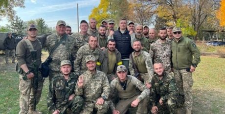 A group of people in military uniforms and casual clothing pose together outdoors, surrounded by trees with autumn foliage.