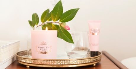 A lit candle, a leafy branch, a glass, and a pink tube on an ornate tray atop a wooden surface.