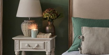 A bedside table with a lamp, a lit candle, a book, and a vase with flowers. The bed has green and beige pillows against a green wall.