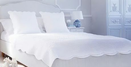 A neatly made bed with white pillows and a scalloped-edge blanket in a bright, white bedroom with a bedside lamp and decorative cabinet.