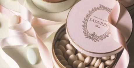 Open Ladurée box with sugared almonds, surrounded by pink ribbons on a light surface.