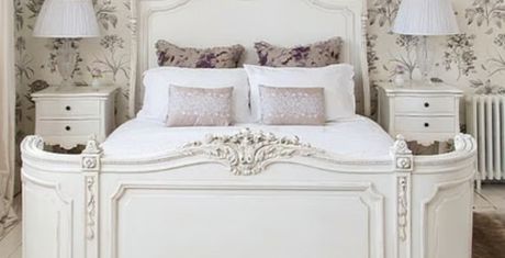 Elegant white vintage bed with ornate carvings, flanked by matching nightstands and lamps, set against floral wallpaper.