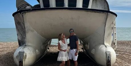 A couple stands under a beached catamaran on a pebble beach, smiling at each other. The sea is visible in the background.