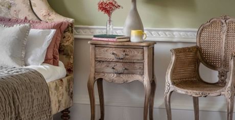 Cozy bedroom with a floral bed, vintage nightstand, books, a lamp, and a wicker chair against a pale green wall.