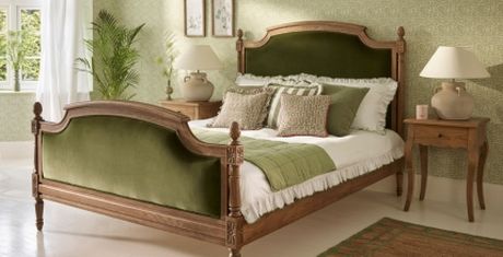 Elegant bedroom with a wooden bed frame, green velvet headboard, matching pillows, and a nightstand with a lamp. Green wallpaper and plants accentuate the decor.