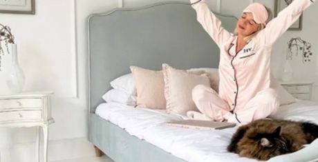 Person in pink pajamas stretching on a bed with a fluffy cat lying nearby. The room is bright and decorated in white and pastel tones.