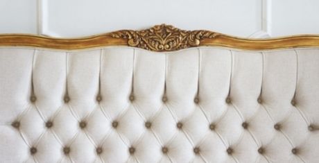 Ornate beige tufted headboard with intricate gold detailing at the top, set against a white wall.
