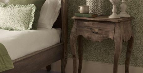 Rustic bedroom scene with a wooden bed, green and white pillows, and a vintage nightstand holding a book, cup, and decorative items.