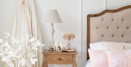 Elegant bedroom with a tufted headboard, wooden nightstand, lamp, white flowers, and pink pillows. Neutral tones create a serene atmosphere.