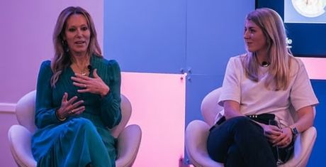 Two women seated on stage, engaged in conversation. One wears a green dress, the other a white top. Purple and pink lighting in the background.