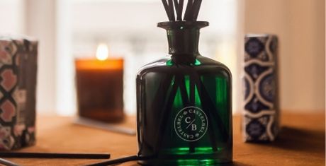 Green glass diffuser bottle with black reeds, surrounded by decorative boxes and a lit candle in the background on a wooden surface.