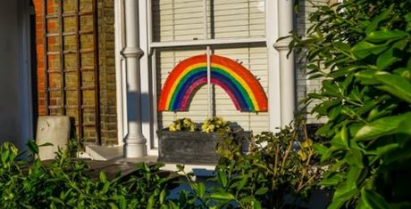 A colorful rainbow drawing displayed in a window with white blinds, surrounded by green foliage and a flower box beneath it.