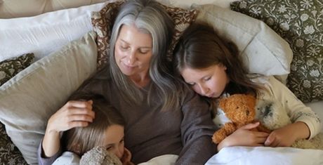 A woman with gray hair cuddles two children in bed, each holding a teddy bear, surrounded by patterned pillows.
