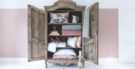 An open vintage wooden wardrobe displaying folded blankets, pillows, books, a white pitcher, and decorative items against a pink and white wall.