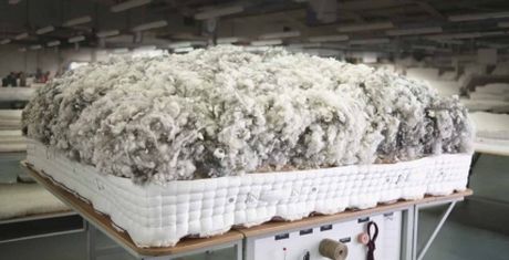 A large, fluffy mattress with exposed stuffing sits on a table in a factory setting, showcasing its inner materials.