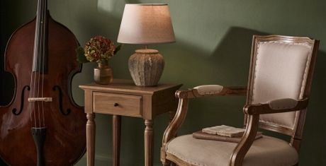 Cozy room with a wooden chair, side table with a lamp and flowers, and a double bass against a green wall.