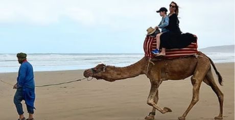 A person leads a camel on a beach, with two people seated on the camel's back, one wearing a hat and the other in a black outfit.