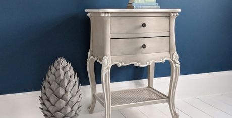 Elegant vintage-style nightstand with two drawers and curved legs, set against a blue wall. A decorative pinecone sculpture is beside it.