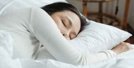 Person sleeping peacefully on a white pillow, wearing a light-colored shirt, with a wooden chair in the background.