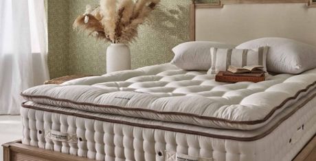 A cozy, well-made bed with a plush mattress and pillows, a striped cushion, and a book. A vase with pampas grass is in the background.