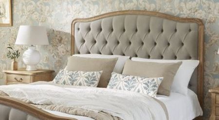 Elegant bedroom with a tufted headboard, patterned wallpaper, and a neatly made bed with decorative pillows and a white lamp on a wooden nightstand.